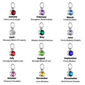 May include: A collection of twelve silver-toned metal charms, each featuring a circular gemstone in a unique color representing a month. The text below each charm lists the month and associated traits like Faith, Love, Courage, and Good fortune.