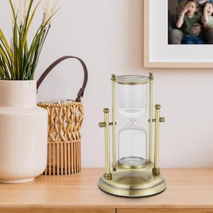 Op de afbeelding: Een zandloper van goudkleurig metaal met een glazen bol en een metalen standaard. De zandloper staat op een houten oppervlak.