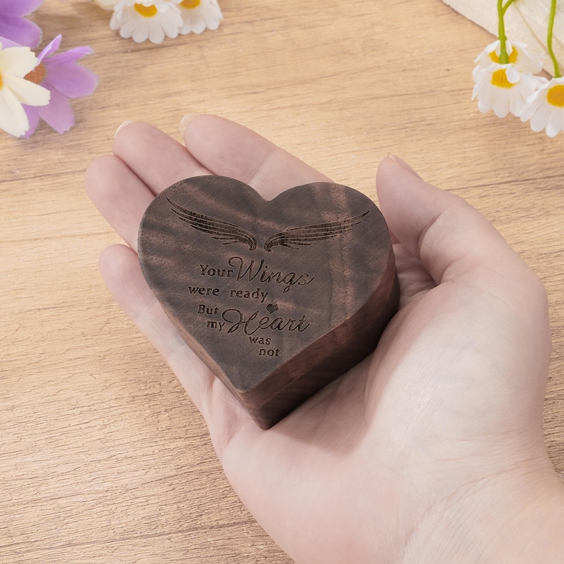 May include: A heart-shaped wooden box with engraved text and wing design. The box is dark brown and held in a hand. The text reads "Your Wings were ready But my Heart was not."
