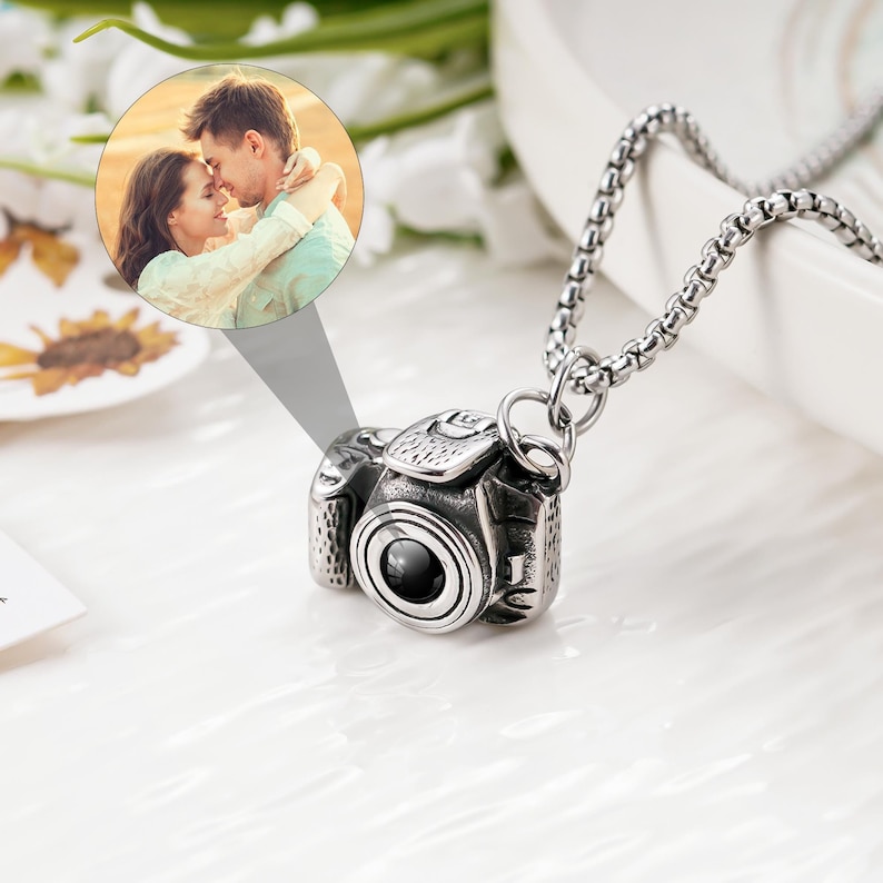 May include: A silver-toned metal camera pendant necklace featuring a detailed miniature camera charm with a central lens. A circular projection insert displays a photograph of a couple embracing. The pendant hangs from a silver-toned box chain.