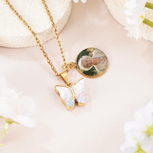 May include: A gold-toned chain necklace featuring a butterfly-shaped pendant with an iridescent white inlay and a circular charm containing a photographic portrait of an older person with white hair wearing a beige garment.