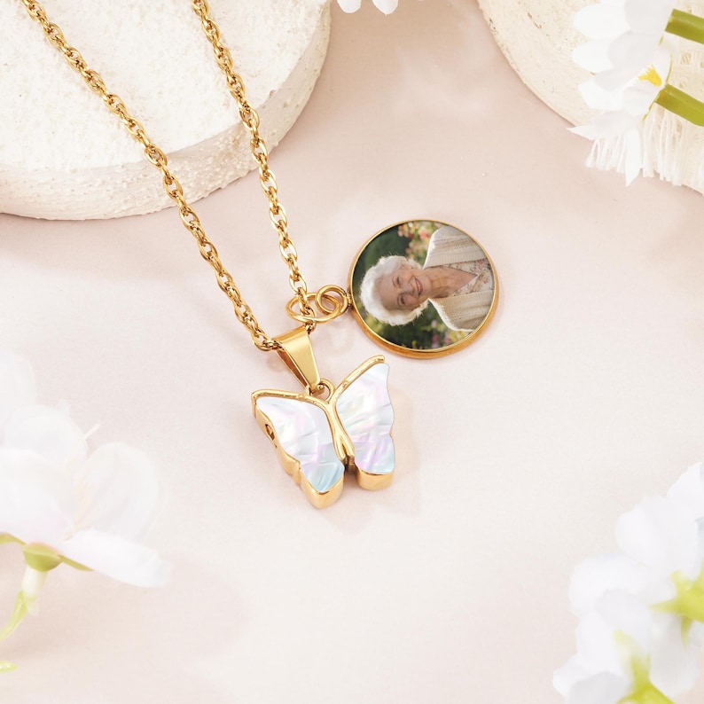 May include: A gold-toned chain necklace featuring a butterfly-shaped pendant with an iridescent white inlay and a circular charm containing a photographic portrait of an older person with white hair wearing a beige garment.