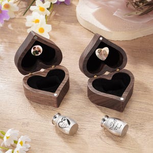 May include: Two open, heart-shaped wooden boxes with a dark interior and a small photo inside. Two small silver urns are also present. The boxes and urns are on a light wood surface, with flowers in the background.