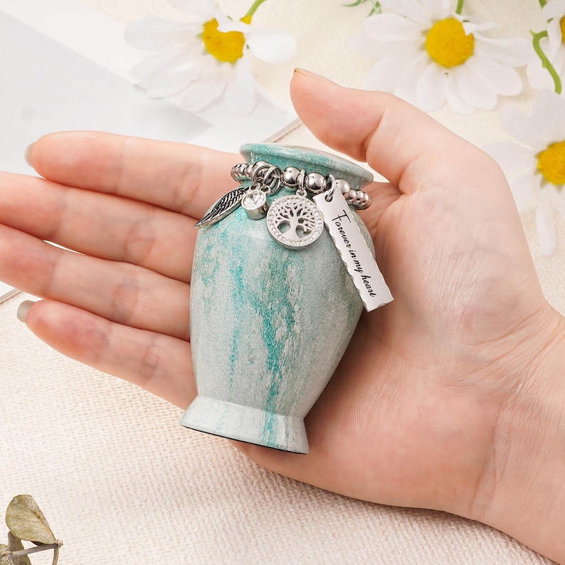 May include: A small, teal and white marbled keepsake urn featuring a silver-toned beaded chain around the neck. The chain holds three charms: a small angel wing, a circular tree of life pendant, and a rectangular tag inscribed with Forever in my heart.