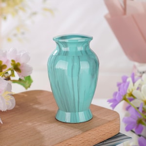 May include: A small, teal-colored urn with a glossy finish and a subtle marbled pattern. The vessel features a classic vase-like shape with a flared rim and a sturdy, circular base, providing a smooth, reflective surface throughout its exterior.