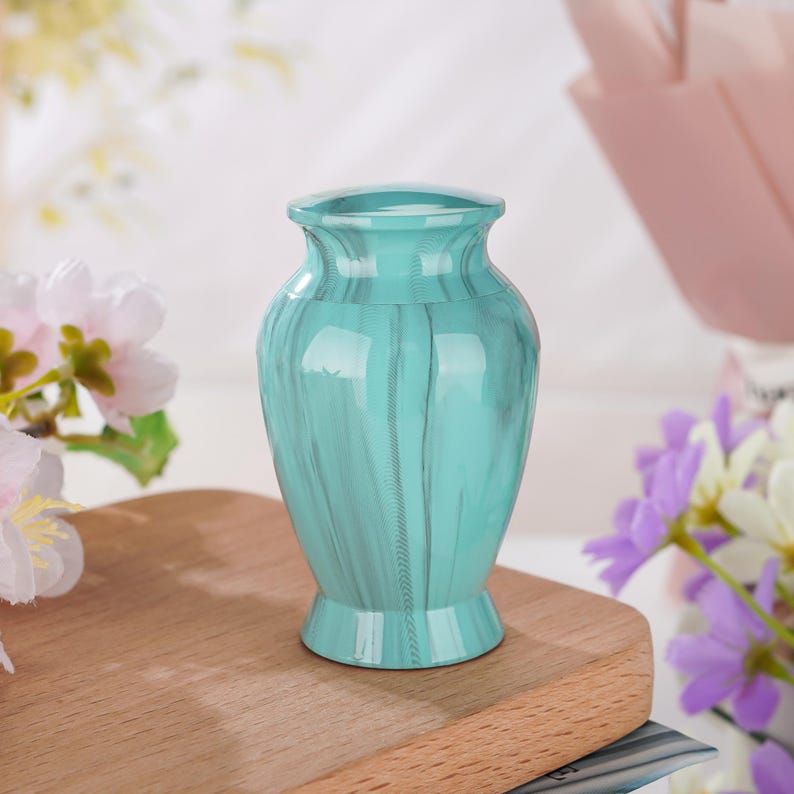 May include: A small, teal-colored urn with a glossy finish and a subtle marbled pattern. The vessel features a classic vase-like shape with a flared rim and a sturdy, circular base, providing a smooth, reflective surface throughout its exterior.
