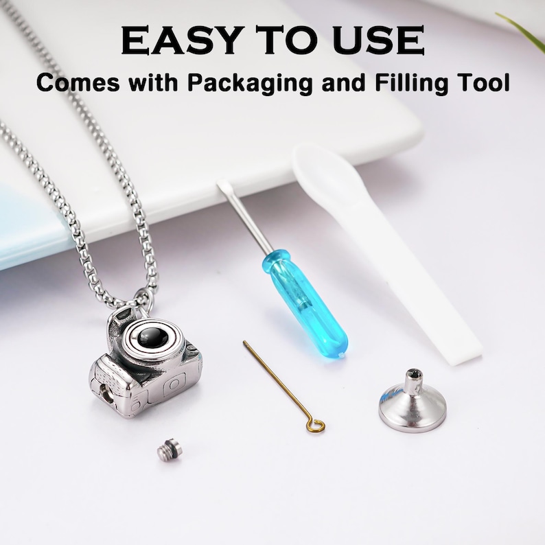 May include: A silver-toned camera pendant necklace with a chain, accompanied by a small screwdriver with a blue handle, a white plastic spatula, a thin metal pin, and a small metal funnel. Text reads: EASY TO USE, Comes with Packaging and Filling Tool.
