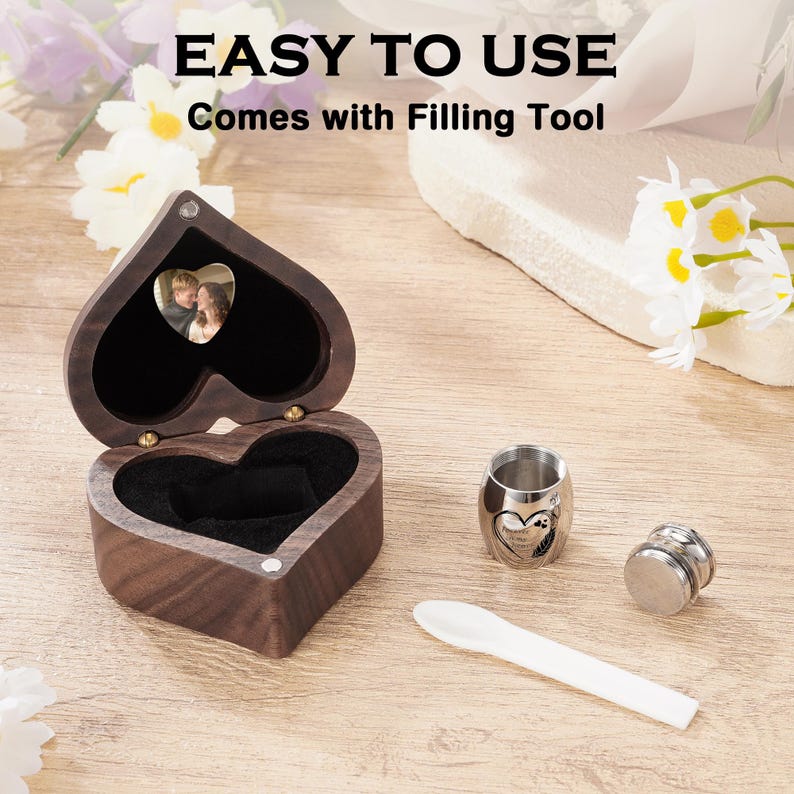 May include: A heart-shaped wooden keepsake box with a photo inside, open to reveal a black interior. Includes a small silver urn, a filling tool, and a small silver container. The text "EASY TO USE" and "Comes with Filling Tool" is visible.