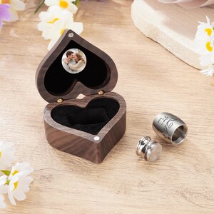 May include: A heart-shaped wooden box with a photo inside, open to reveal a black interior. Next to it are two silver metal containers, one with engraved text. The box is on a wooden surface.
