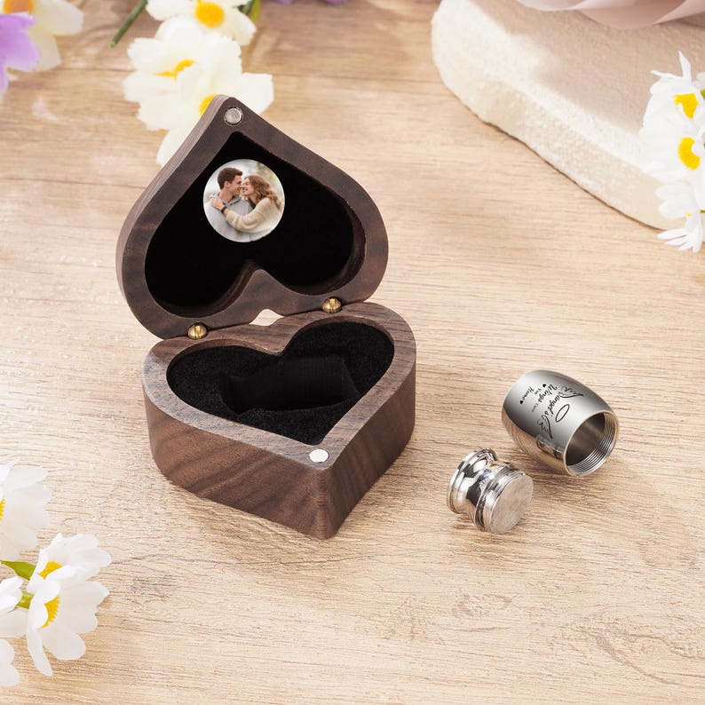 May include: A heart-shaped wooden box with a photo inside, open to reveal a black interior. Next to it are two silver metal containers, one with engraved text. The box is on a wooden surface.