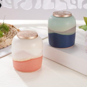 May include: Two ceramic jars with lids. The jar on the left is white with a peach colored bottom and a gold lid. The jar on the right is blue with a green top and a gold lid.
