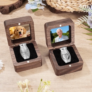 May include: Two wooden keepsake boxes, each with a photo and a small silver urn inside. One box features a photo of a golden retriever, the other a man. The urns have engraved designs and text, suitable for memorial keepsakes.