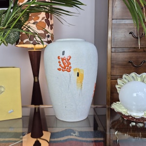 Könnte beinhalten: Eine weiße Keramikvase mit strukturierter Oberfläche und abstrakten Blumendesigns in Orange und Gelb. Die Vase hat eine runde Form und steht auf einer Glasoberfläche.