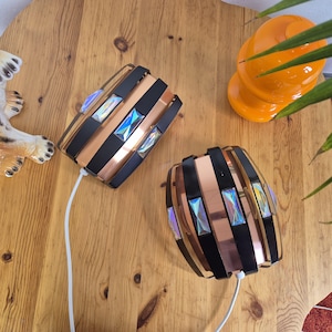 Può includere: Due lampade decorative con un design rame e nero. Ogni lampada presenta accenti rettangolari iridescenti e un cavo di alimentazione bianco. Le lampade sono cilindriche e poggiano su una superficie di legno.