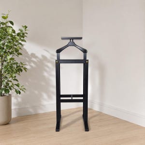May include: A black wooden valet stand featuring a top hanger bar for jackets, a flat horizontal shelf, and a rectangular frame base. The structure includes two small metallic gold-toned circular accents on the front frame joints.