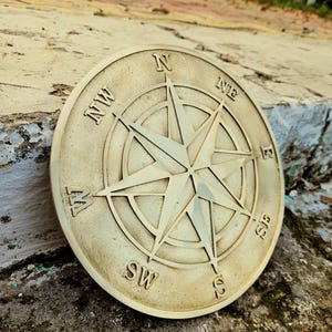 Peut inclure: Une plaque circulaire en métal doré représentant une rose des vents avec un motif d'étoile centrale et des points cardinaux et intercardinaux en relief, incluant N, NE, E, SE, S, SW, W et NW gravés sur le pourtour du cadran.