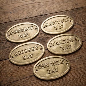 Könnte beinhalten: Fünf ovale Messingschilder mit erhabener Schrift. Die Schilder tragen die Aufschriften PORTAGE BAY, HAYSTACK BAY, WHISKEY BAY, TRADING BAY und TEN MILE BAY. Jedes Schild hat zwei kleine Löcher zur Befestigung, eines links und eines rechts.