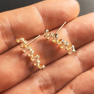 May include: A pair of gold earrings with a row of clear, marquise-shaped stones. The earrings are curved and have a delicate, minimalist design.