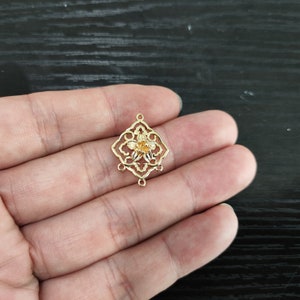 May include: A gold-colored filigree charm with a floral design. The charm has a diamond-shaped frame with an openwork pattern and a small flower in the center. It has four loops for attaching to other jewelry components.