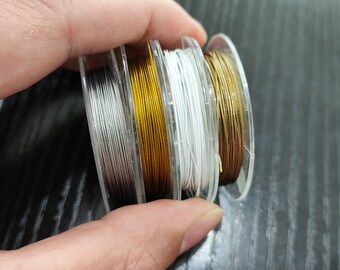 May include: Three spools of thread, one silver, one gold, and one white, are stacked on top of each other. The spools are held in a hand.