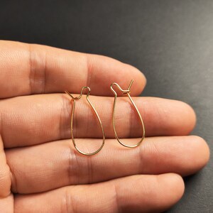 May include: A pair of gold-toned earring hooks, also known as ear wires, are shown on a hand. The hooks are made of thin, flexible wire and have a teardrop shape.