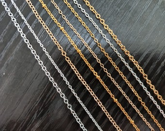 Könnte beinhalten: Eine Sammlung von silbernen und goldenen Ketten in verschiedenen Längen und Stärken. Die Ketten sind diagonal auf einer dunklen Holzoberfläche angeordnet.