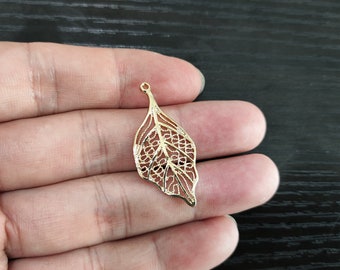May include: A gold-colored metal leaf charm with a delicate, intricate design. The charm has a loop at the top for attaching to jewelry or other crafts.