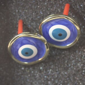 May include: Two gold-toned cufflinks with blue evil eye charms. The charms have a white circle with a blue pupil and a black dot in the center.