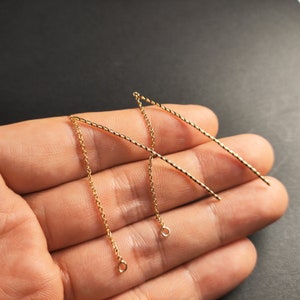 Może przedstawiać: Dwa złote kolczyki z łańcuszka o splecionym wzorze. Kolczyki wiszą z dłoni.