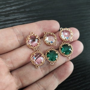 May include: Six gold-tone connector charms with different coloured glass stones. The charms feature a rope-like design around the edges. The stones are pink, clear, green, and a darker green.