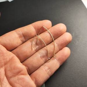 Puede incluir: Dos ganchos para pendientes de color dorado, también conocidos como alambres para orejas, se muestran en una mano. Los ganchos están hechos de metal delgado y tienen un diseño simple y curvo.