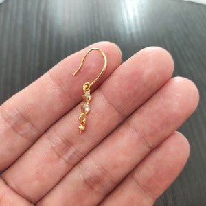 Könnte beinhalten: Ein goldener Ohrring mit einem kleinen, klaren Kristall-Anhänger. Der Ohrring hat ein schlichtes, elegantes Design.