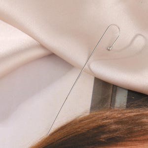 May include: A single, silver-colored ear cuff earring. The earring has a long, straight bar that curves at the top, ending in a small, round bead. The earring is set against a backdrop of a light pink fabric.