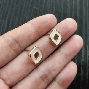 May include: Two gold-toned square-shaped earring blanks with a white center and a small loop on the top.