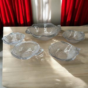 May include: A collection of five clear glass serving bowls, each with a seashell pattern. The bowls are of varying sizes, with a large central bowl and smaller ones around it. They are displayed on a light-coloured wooden surface.