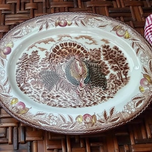May include: Oval serving platter with a turkey and forest scene in brown tones. The rim features a decorative border of fruits and foliage. A red and white checkered cloth is visible on the right side.