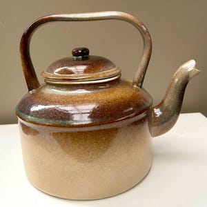 May include: A ceramic teapot featuring a glossy glaze with a gradient transition from a light beige base to a deep brown and green upper section. The teapot has a rounded body, a curved spout, a matching lid with a small knob, and a high arched handle.