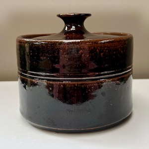 May include: A cylindrical ceramic container with a matching lid, featuring a dark brown, glossy glaze with subtle black and reddish-brown speckling. The lid has a central, flared knob handle, and the overall finish is smooth and reflective.