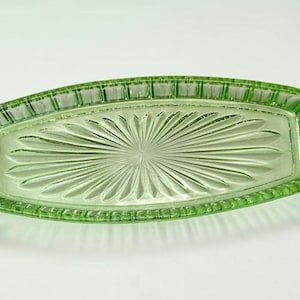 May include: A rectangular, light green glass dish with a sunburst pattern in the center. The edges of the dish have a textured, ribbed design. The dish is empty and sits on a white surface.