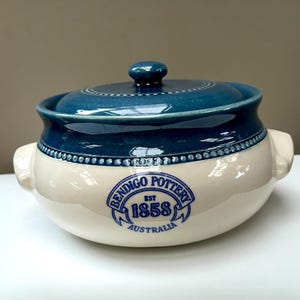 May include: A cream-coloured ceramic casserole dish with a dark blue glazed lid and rim. The body features a blue stamped logo that reads BENDIGO POTTERY EST 1858 AUSTRALIA. The pot has two side handles and a small knob on the lid.