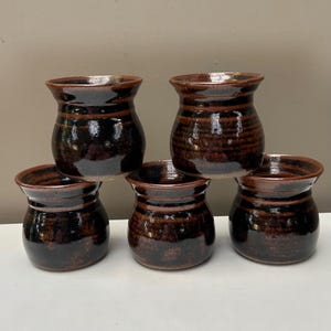 May include: A set of five small, rounded ceramic cups with a glossy, dark brown glaze. Each cup features a flared rim and a bulbous body with subtle horizontal ridges, showcasing a consistent, reflective finish across all five individual pieces.