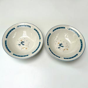 May include: Two off-white ceramic bowls featuring a central illustration of a white goose wearing a blue polka-dot ribbon. The rim of each bowl is decorated with a dark teal band containing small white floral accents and a thin blue line.