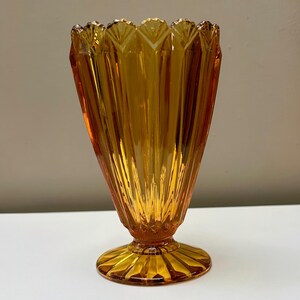 May include: Amber glass vase with a flared, fan-shaped top and a pedestal base. The vase features vertical ribbed detailing and a scalloped rim. The glass has a warm, golden hue. The vase is likely vintage.