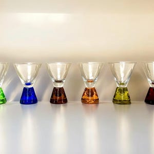 May include: Six clear glass cocktail glasses with colorful bases in green, blue, brown, orange, yellow, and red. The glasses have a conical bowl shape and a triangular base, arranged in a row.