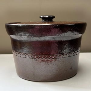 May include: A dark brown ceramic crock with a matching lid. The cylindrical vessel features a textured, metallic-like finish and a horizontal band of raised circular bead detailing encircling the upper portion of the body below the rim.