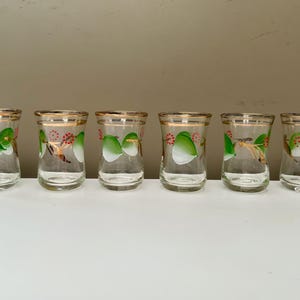 May include: Six clear glass tumblers with gold rims and painted designs. Each glass features a green and white leaf design with red accents. The glasses are arranged in a row on a white surface.