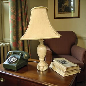 May include: A cream-coloured lampshade sits atop a white marble lamp base on a wooden desk. A dark green rotary phone and a stack of books are also on the desk. A burgundy armchair is visible in the background.