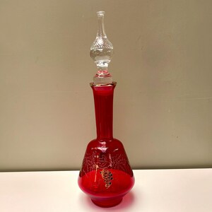 May include: A tall, vibrant red glass decanter featuring a slender neck and a wide, rounded base. The body is decorated with etched gold-coloured grape vine patterns. It includes a clear, textured glass stopper with a bulbous, faceted design.