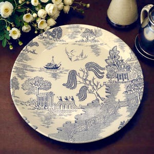 May include: A cream-coloured plate featuring a blue and white pastoral design with boats, trees, and birds. The plate is surrounded by white flowers and rests on a dark wooden surface. The plate's diameter is approximately 25 cm.