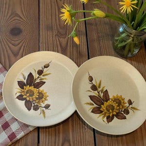 May include: Two beige ceramic plates featuring a sunflower and foliage design. The plates are on a wooden table, with a checkered napkin and a vase of yellow flowers in the background.